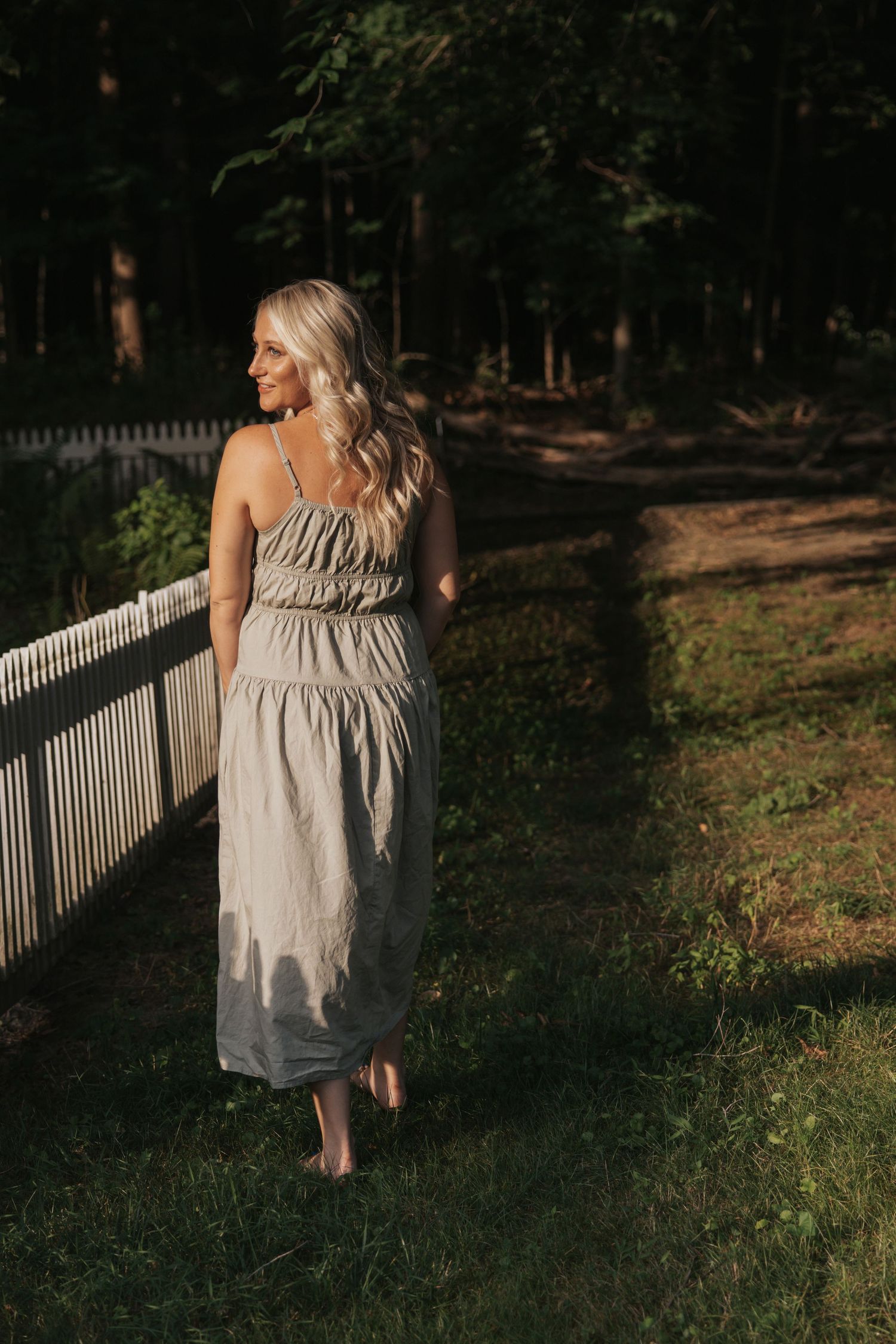 Woman in flowing beige dress walks barefoot along white fence in garden setting.