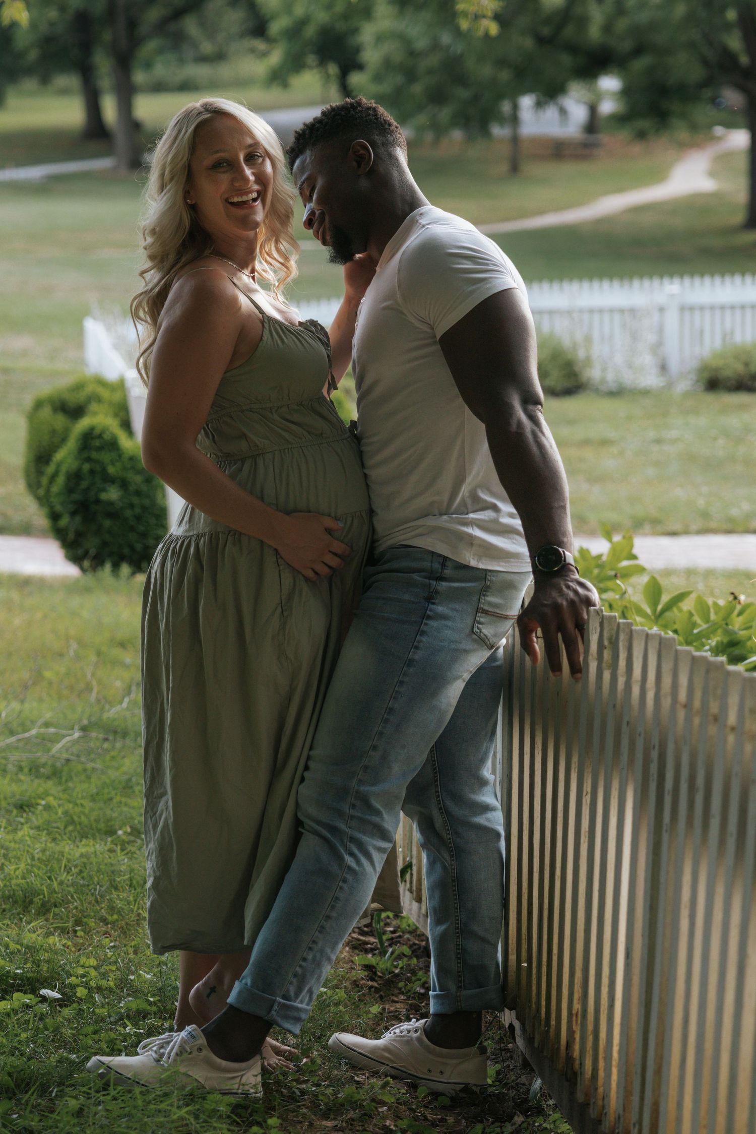 Intimate maternity photo of expectant couple standing by white picket fence in a garden setting.