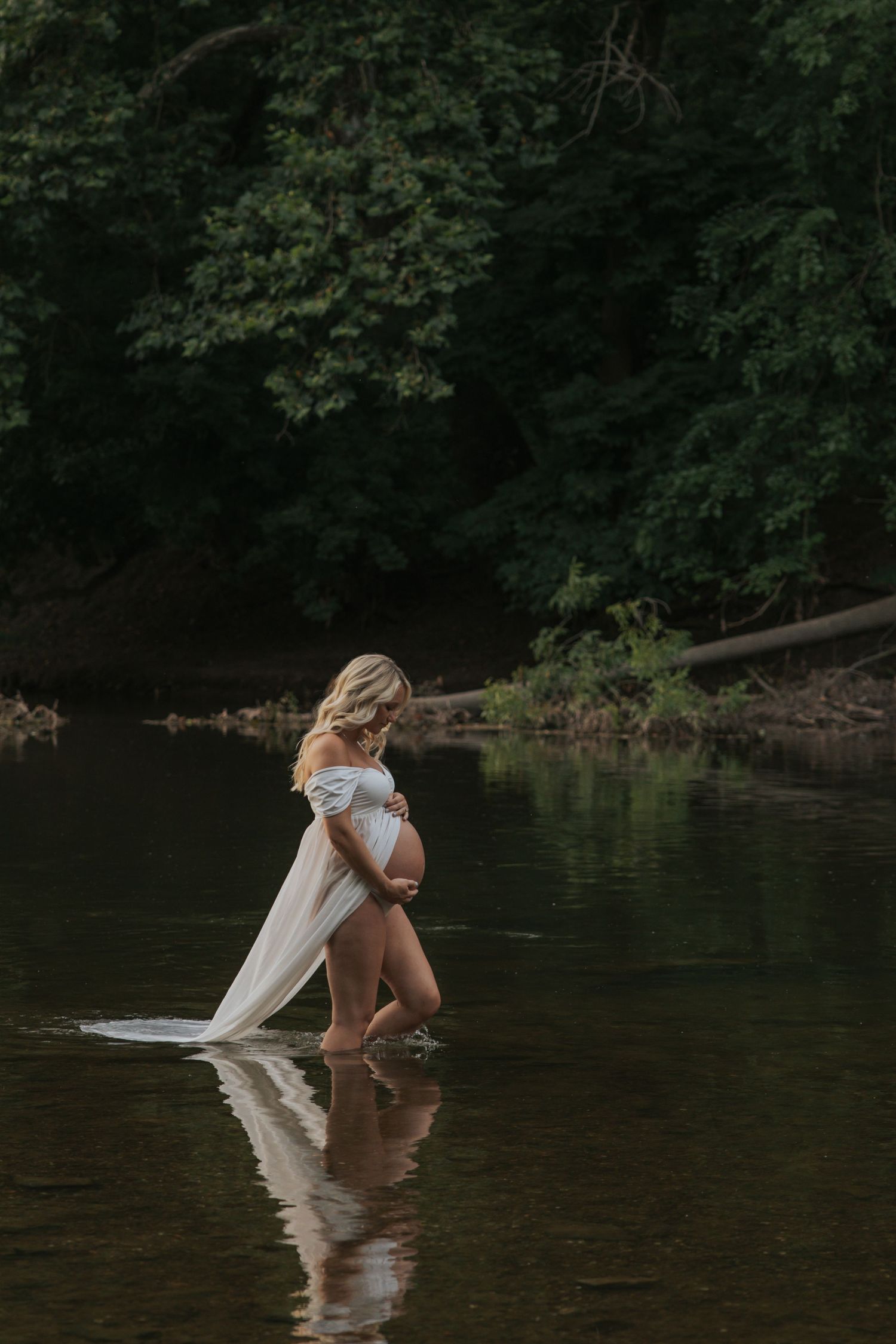 Artistic maternity photo of woman in white dress standing in river with reflection.