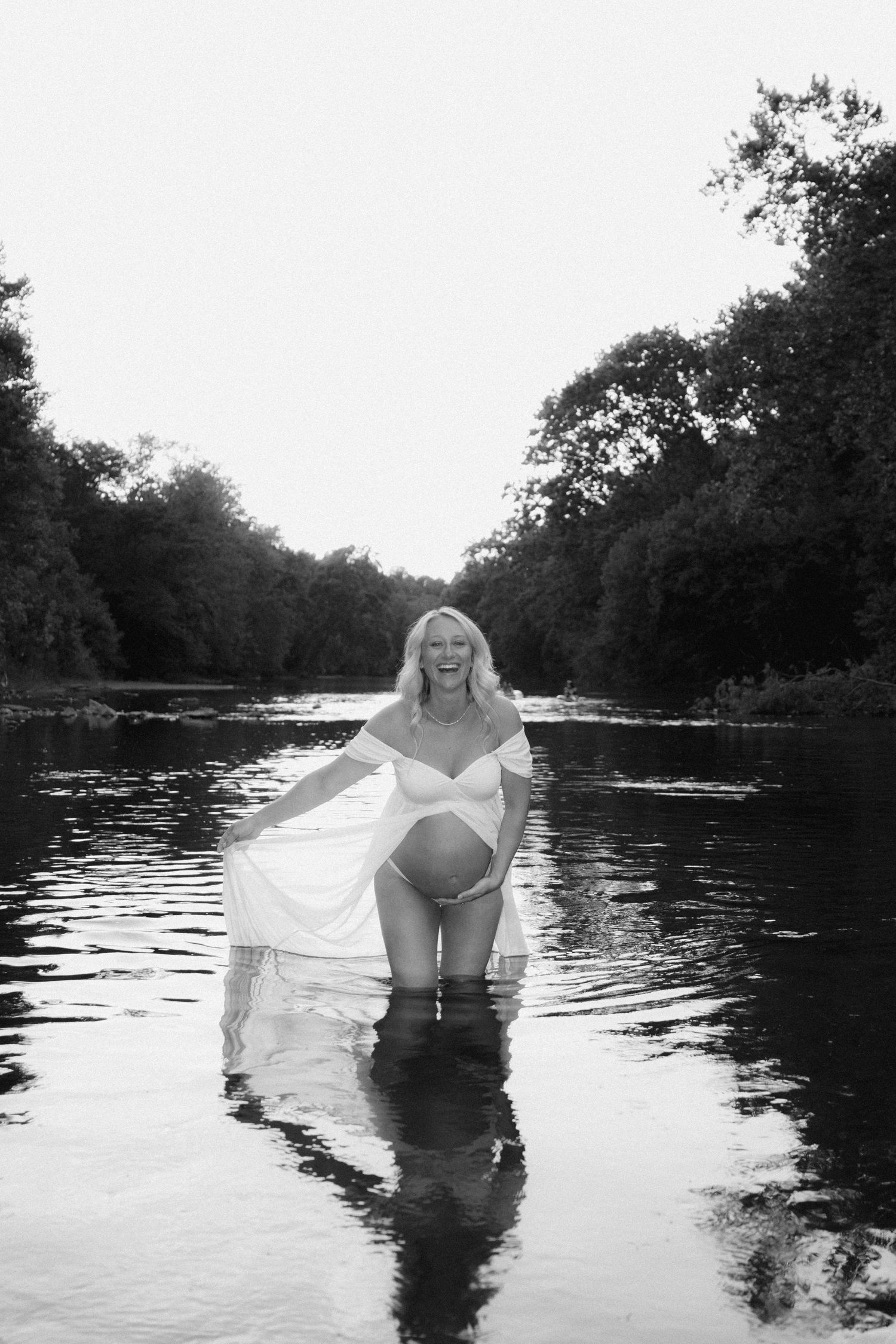 Black and white maternity portrait of woman standing in river wearing flowing dress.