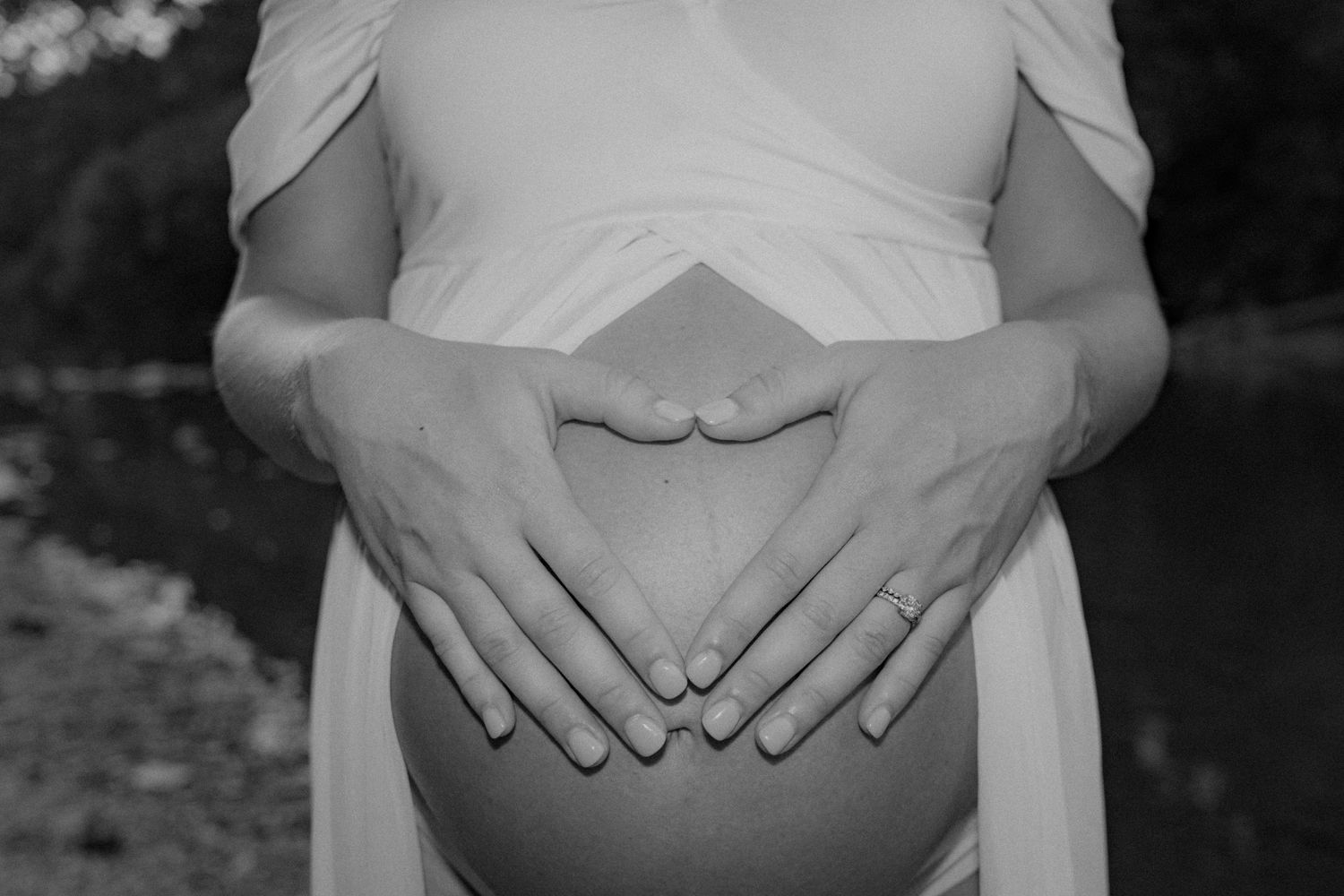 Close-up black and white photo of hands forming heart shape on pregnant belly.