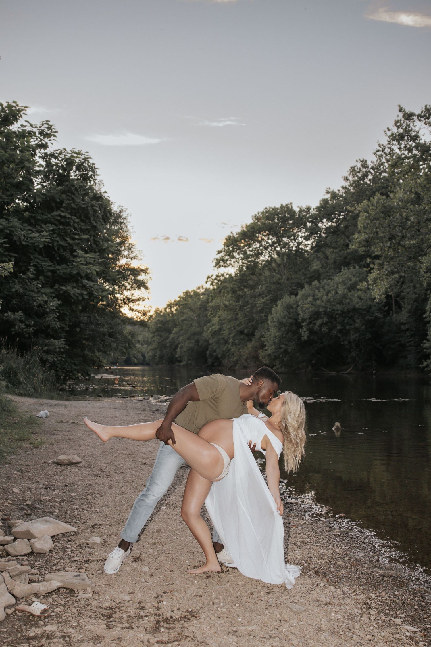 A romantic riverside photoshoot at sunset with a dramatic dip pose along a rocky shoreline.