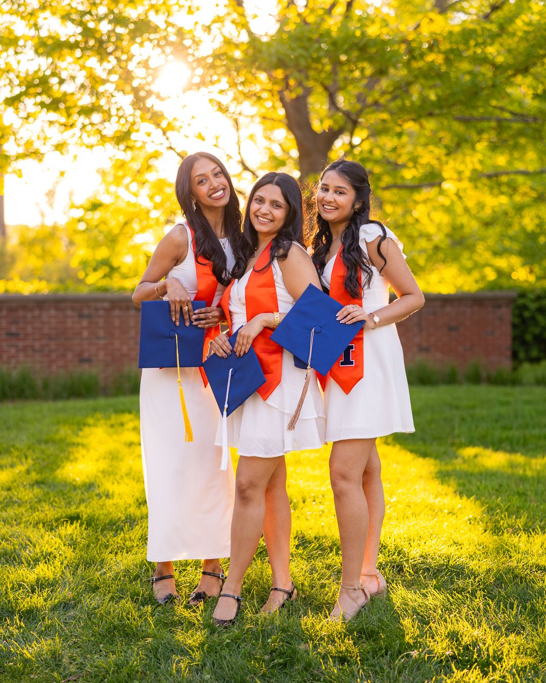 Three girls standing next to each other smiling with UIUC graduation accessories in front of greenery