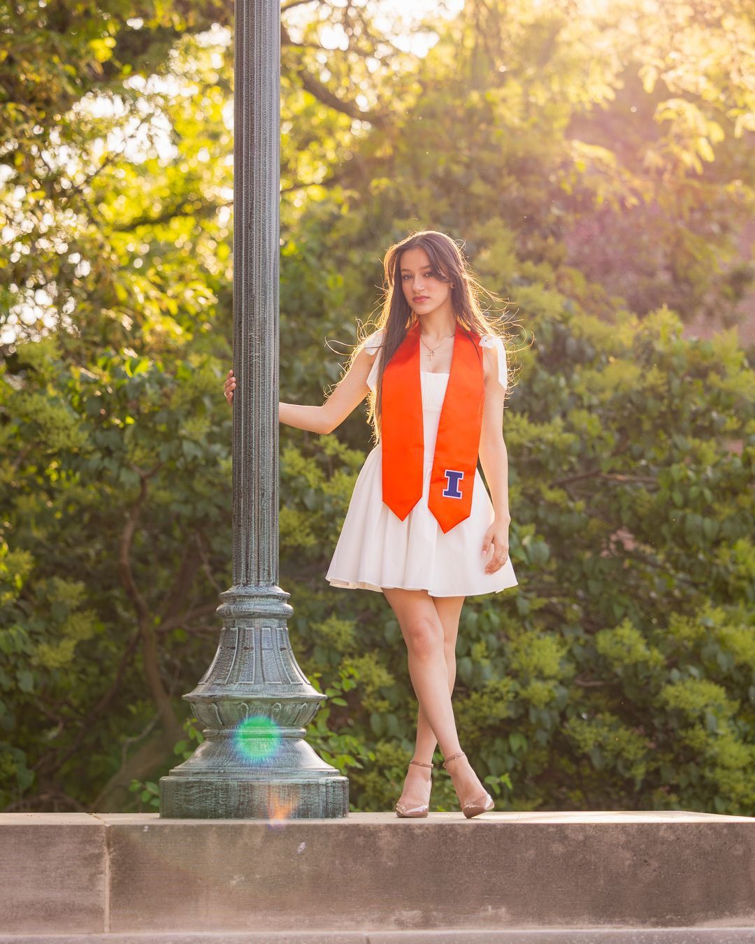 Girl leaning on a pole in front of greenery with graduation attirre at Foellinger Auditorium