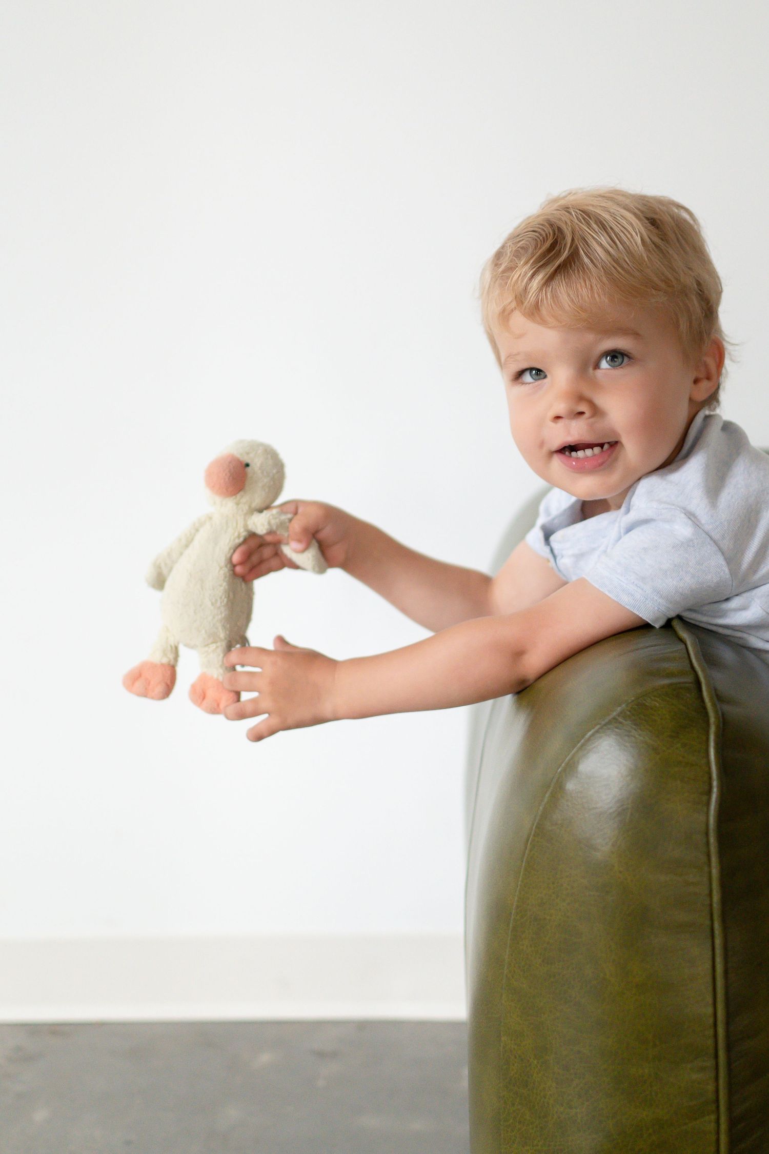 Young child on green leather chair holding small plush toy against white wall.