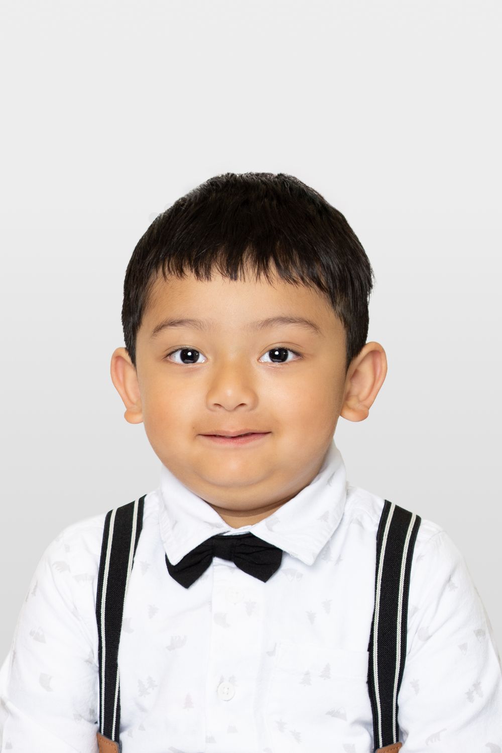 Young child wearing a white dress shirt with black suspenders and bow tie smiling at the camera.