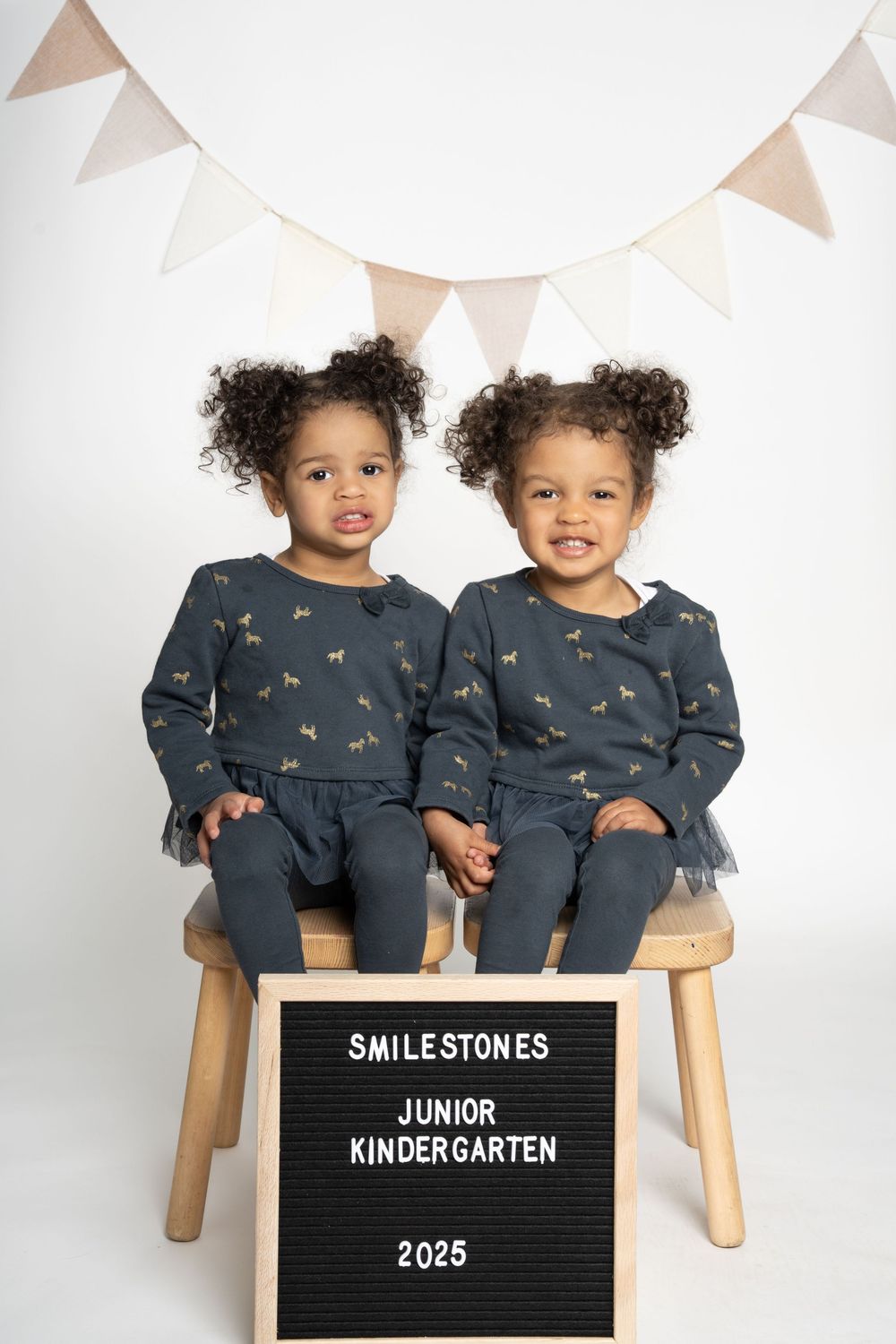 Two children in matching outfits sit on wooden stools with letterboard sign.