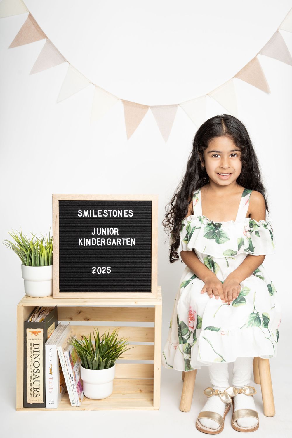 Smiling child in white floral dress sits near letterboard and decorative shelf with plants and bunting.