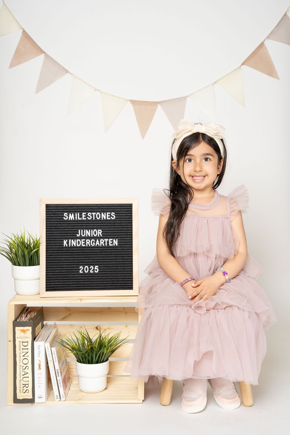 Child in pink tulle dress poses with junior kindergarten milestone board and decorative elements.