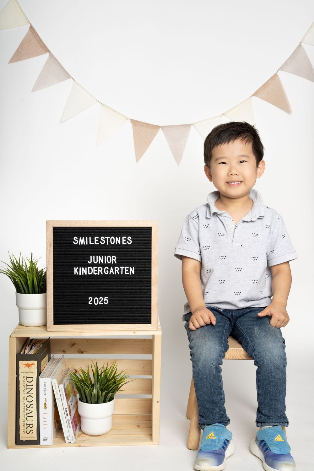 Child in polo shirt and jeans poses with kindergarten milestone board and decorative shelf.
