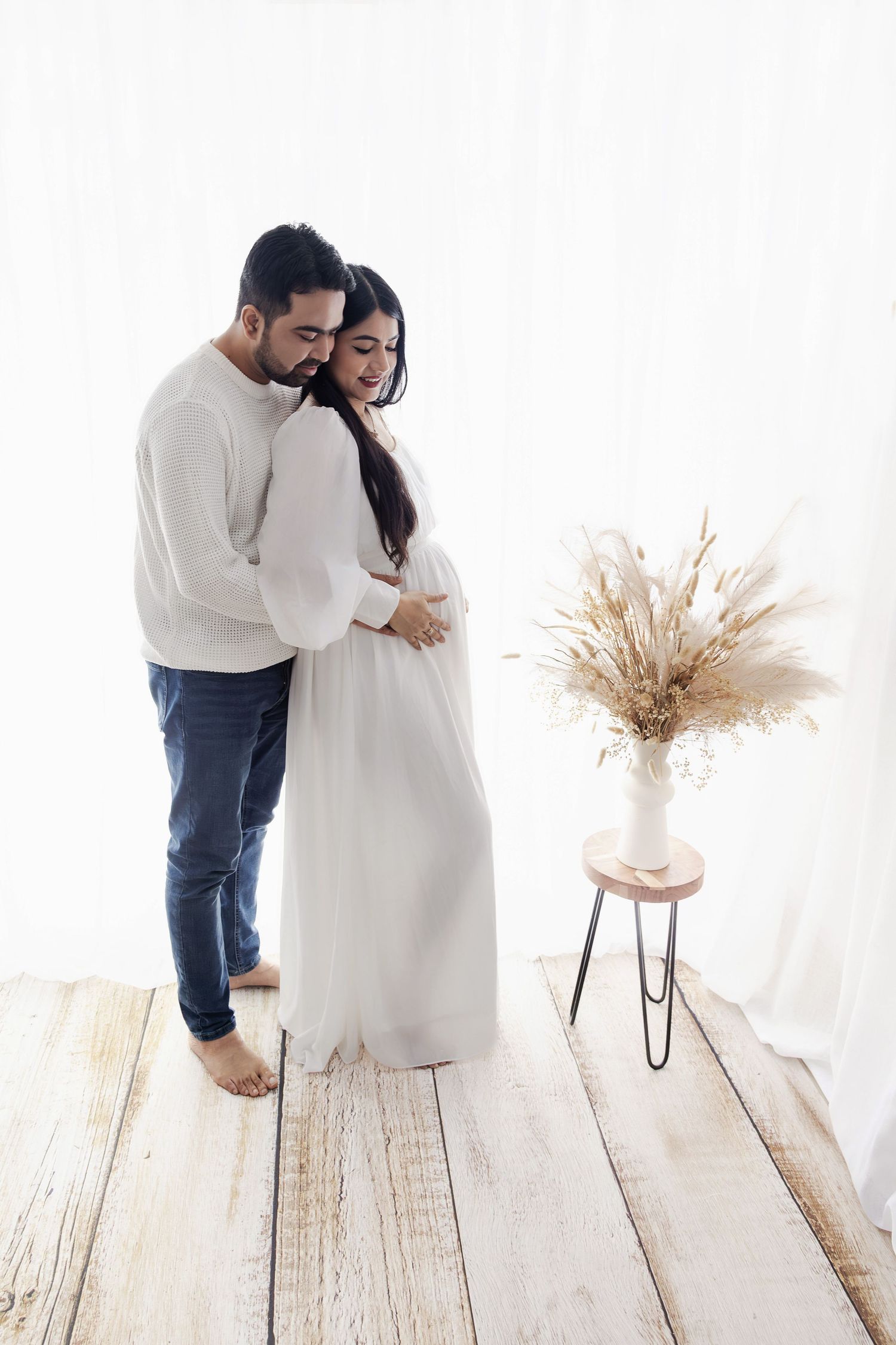 Parents to be standing in my studio with white backlit curtains behind holding hands eagerly anticipating the arrival of their new baby.