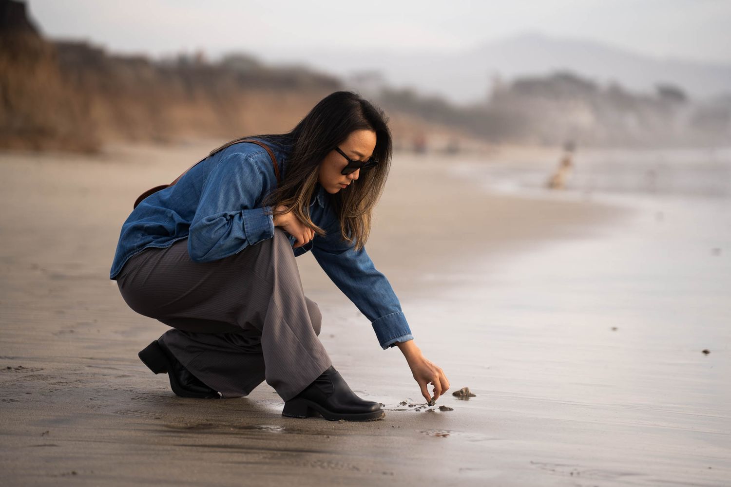 A girl in blue jacket crouches on misty beach writing with a pebble onto the sand at sunset.