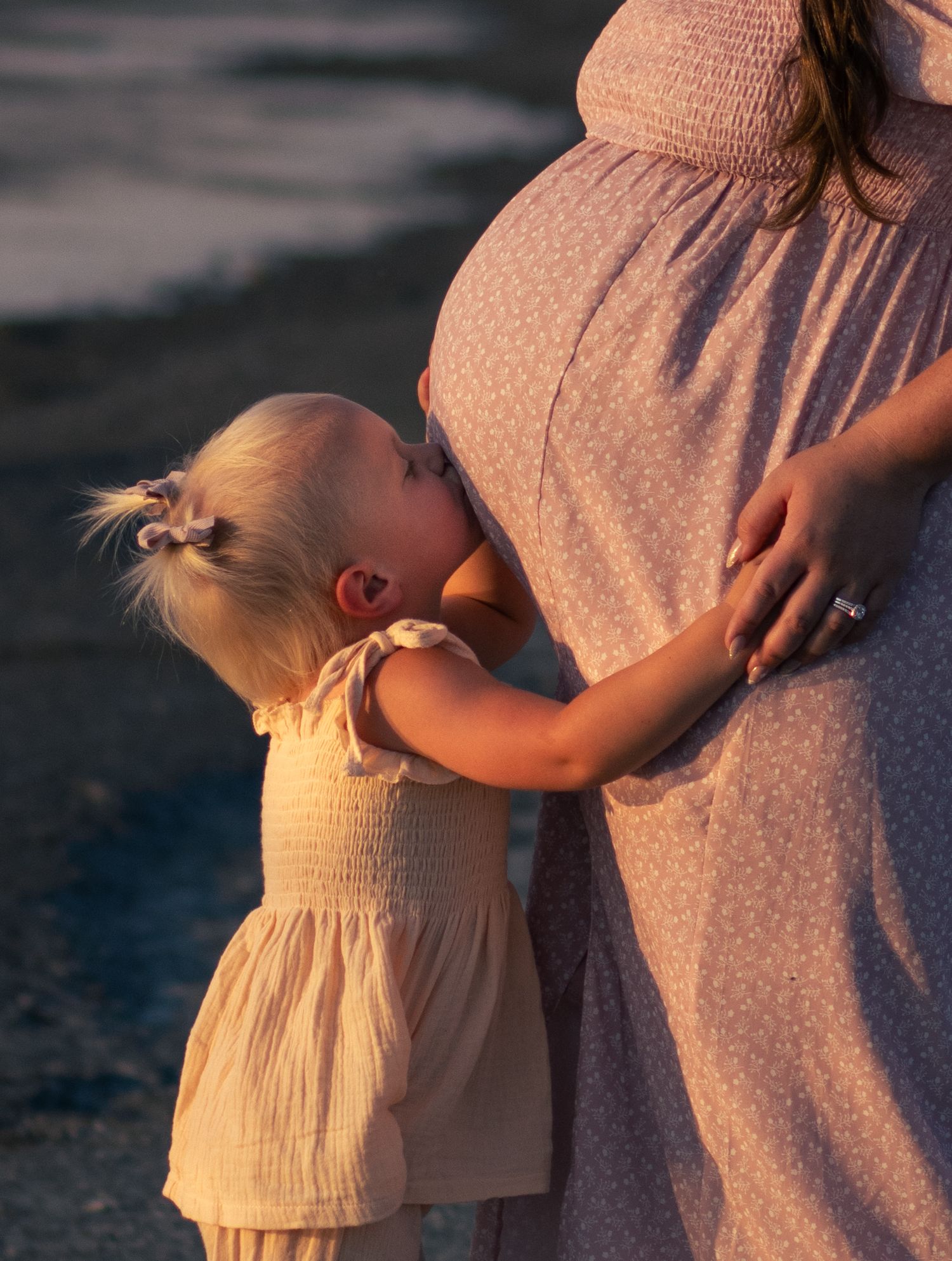 Expecting mother holding baby bump during golden hour maternity session with daughter kissing bump