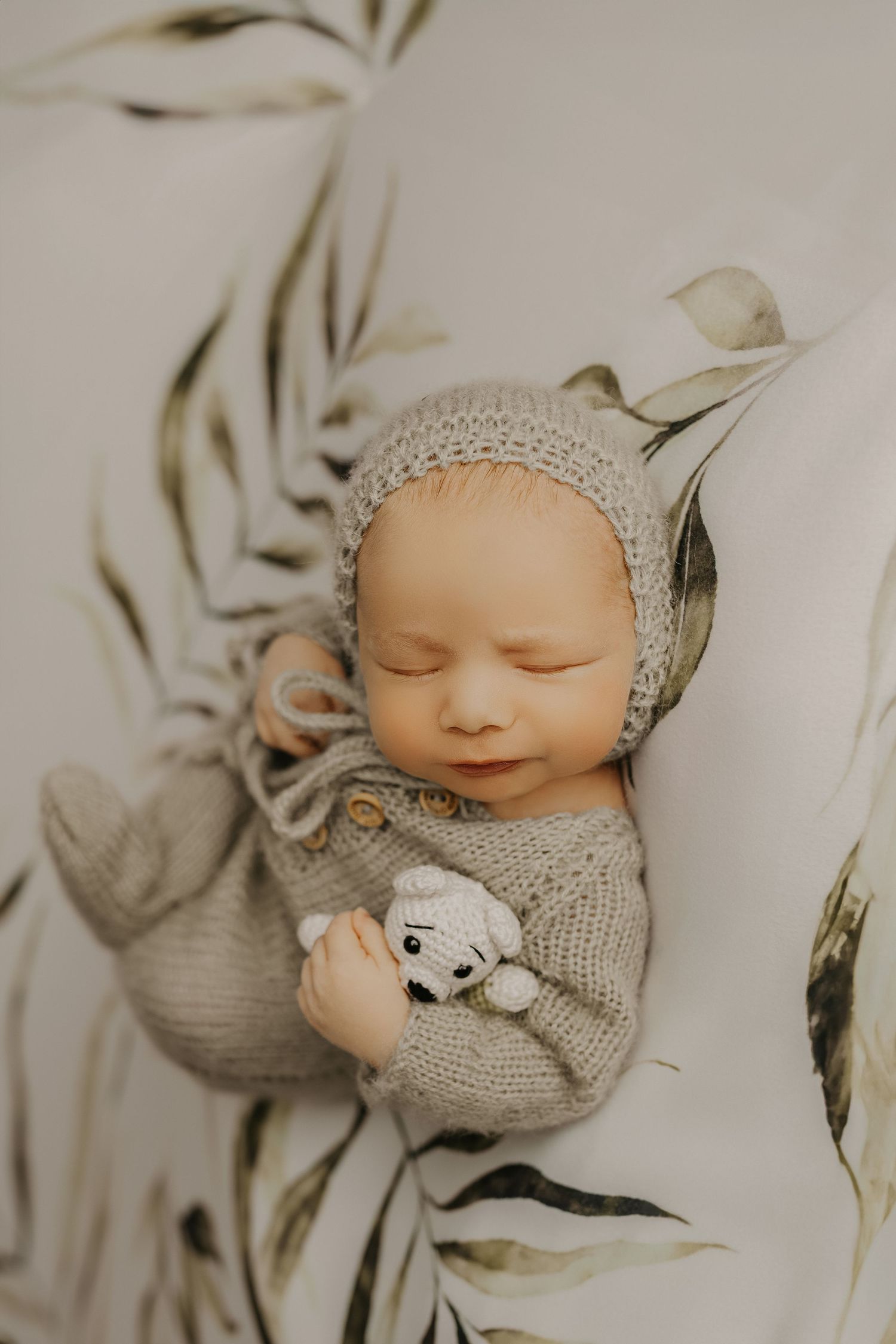 A sleeping newborn baby wearing a gray knit bonnet and sweater lies peacefully on white bedding. Ottawa newborn best photographer near me.