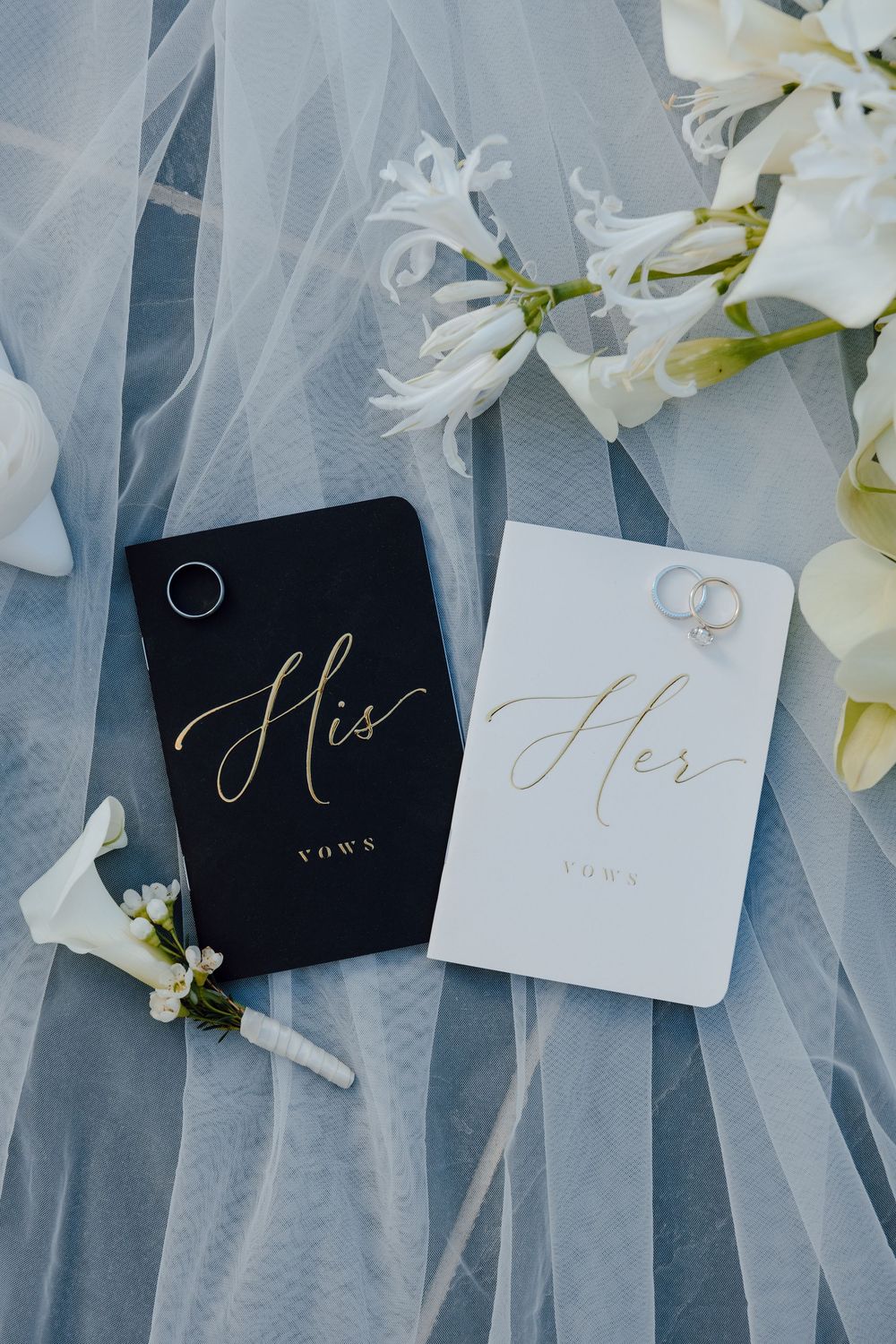 Wedding vow books labeled 'His' and 'Her' with gold lettering displayed on blue tulle fabric with white floral accents.