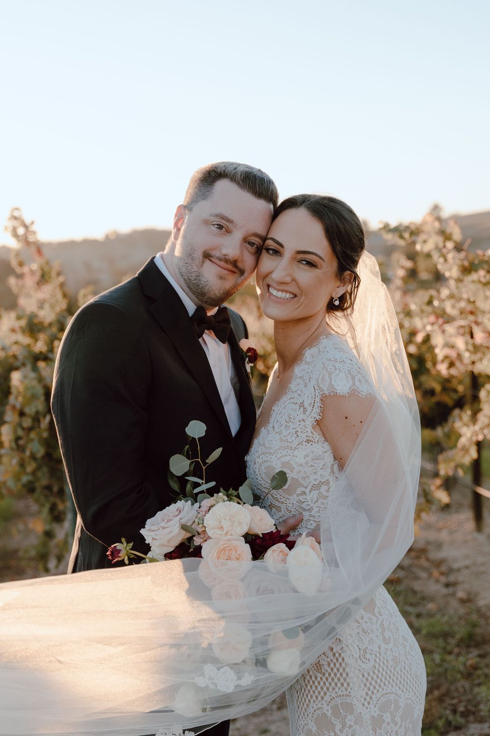 A romantic sunset wedding portrait in a vineyard with delicate flowers and flowing veil.
