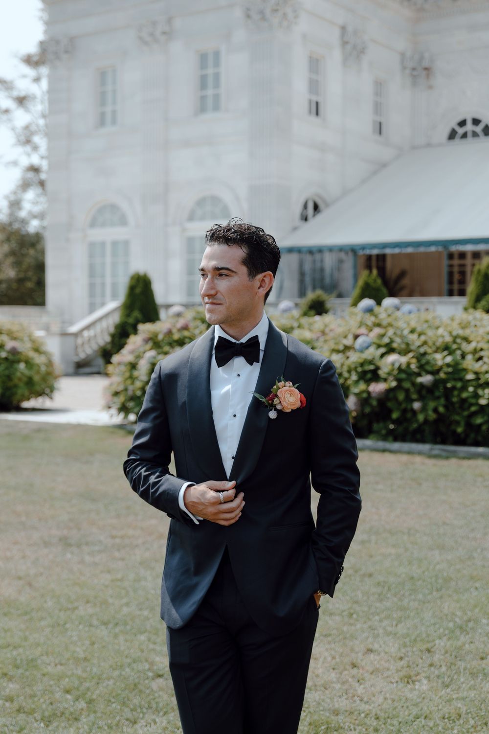 A groom in a classic black tuxedo stands on a manicured lawn in front of a white mansion on his wedding day.
