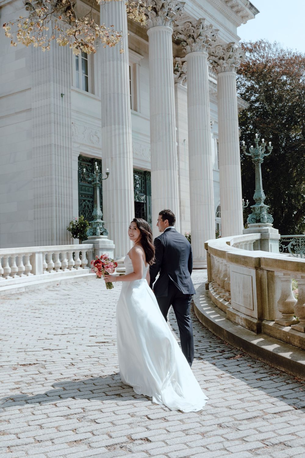 Elegant wedding couple poses on a cobblestone terrace with grand white columns and ornate architecture in the background.