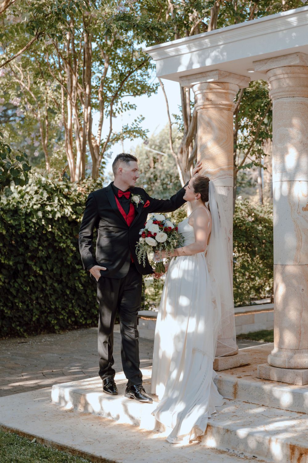 A couple poses under a white columned archway in a garden setting during their wedding ceremony.