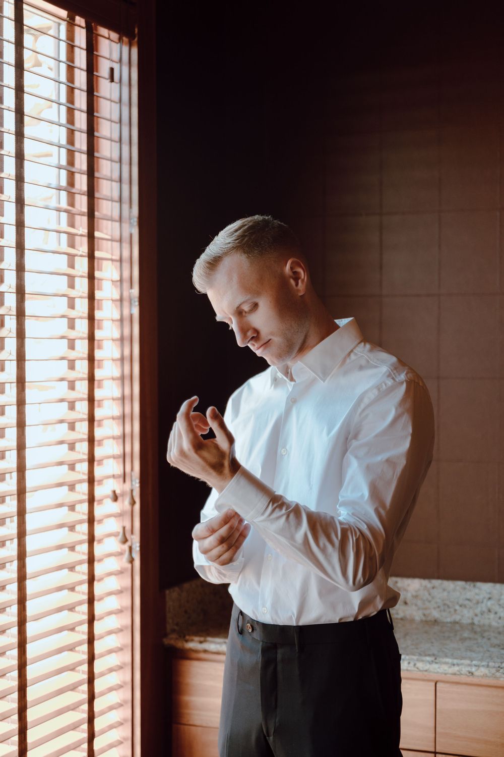 A person in a white dress shirt adjusts their cufflinks by a window with venetian blinds.