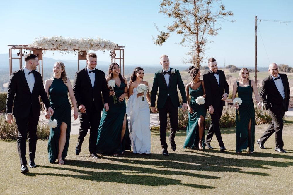 A wedding party walks together on green lawn wearing formal attire and emerald gowns.