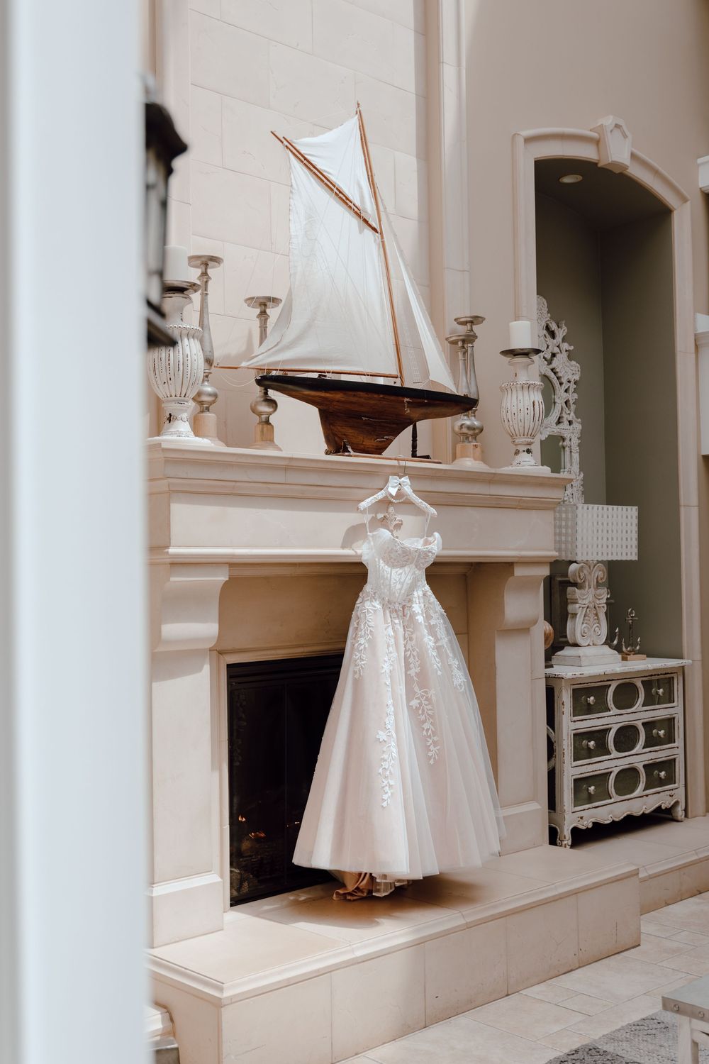 Elegant white lace wedding dress displayed on hanger near fireplace mantel with decorative model sailboat and urns.