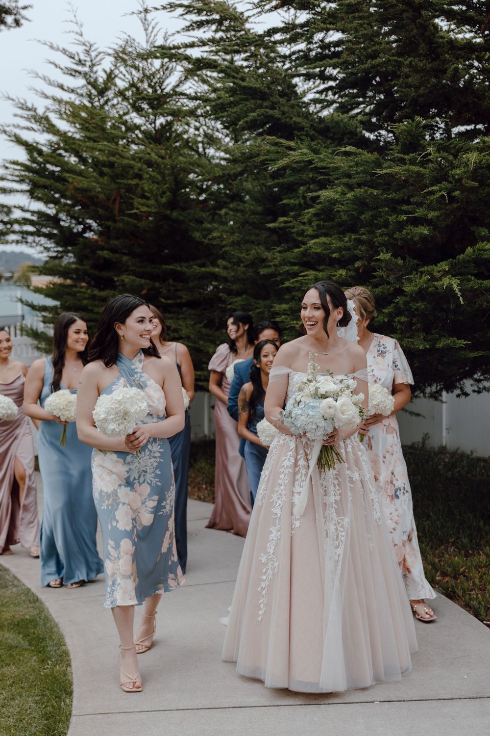 Wedding party in pastel blue and pink dresses gathers outdoors near evergreen trees during a celebration.
