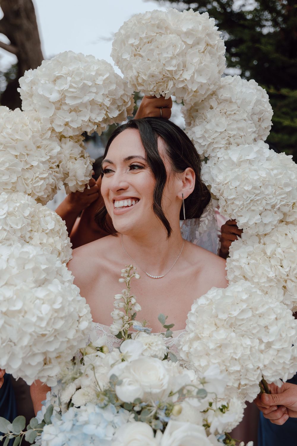 A radiant bride smiles while surrounded by a stunning display of white hydrangea flowers at an outdoor wedding.
