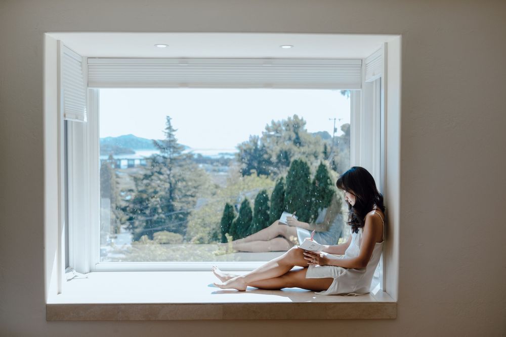 Person lounges on white windowsill overlooking scenic mountain landscape through large window.