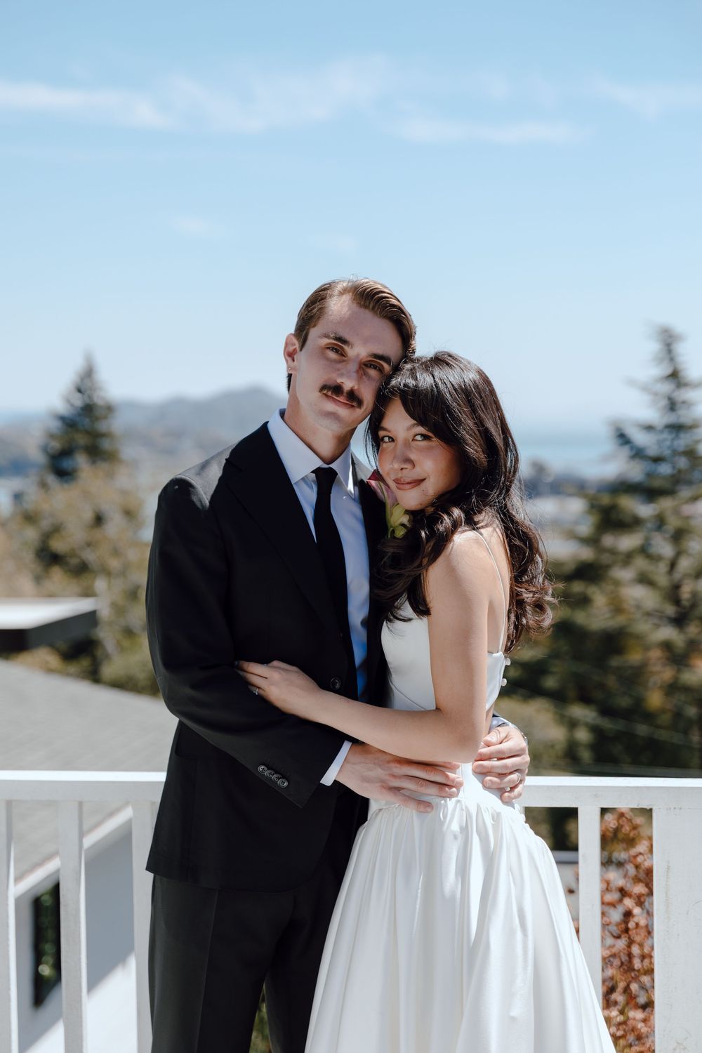 A couple in wedding attire embrace on a balcony overlooking a mountain landscape on their special day.