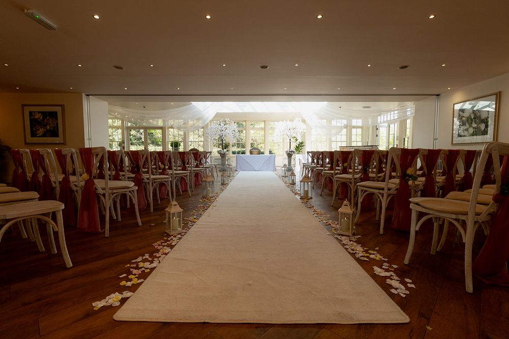 Elegant wedding ceremony aisle with white runner, red chair sashes, and floral decorations in a bright indoor venue.
