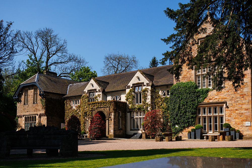 Historic English manor house with ivy-covered brick facade, arched entrance, and manicured grounds under a clear blue sky.