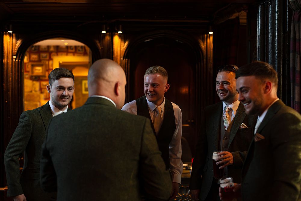 A group of well-dressed men in suits and waistcoats laughing together in a warmly lit elegant venue.