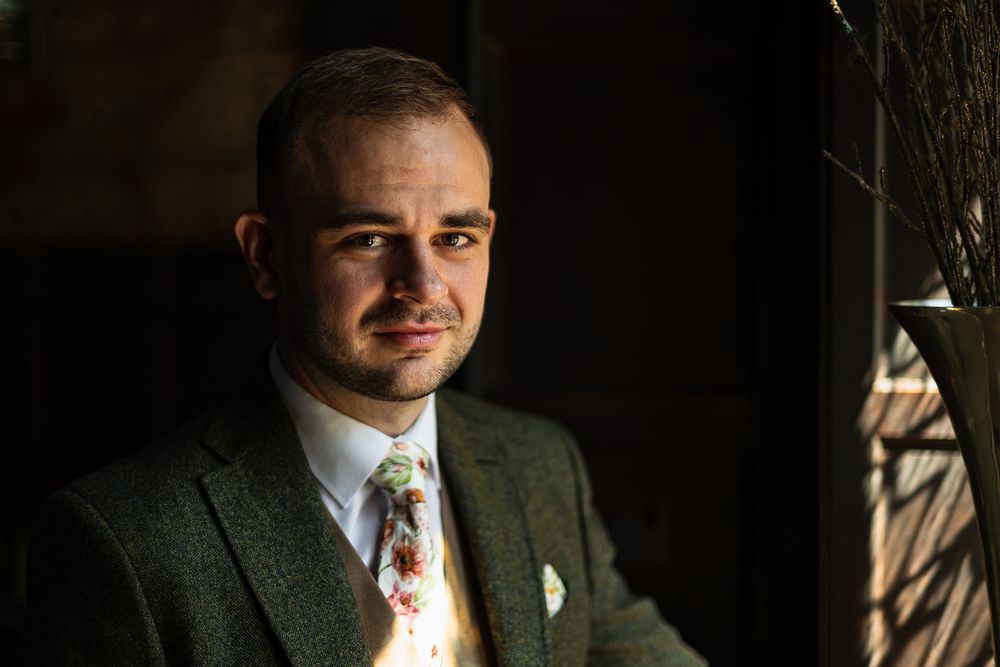 A man in a green tweed suit and floral tie poses thoughtfully near a window in soft, dramatic lighting.