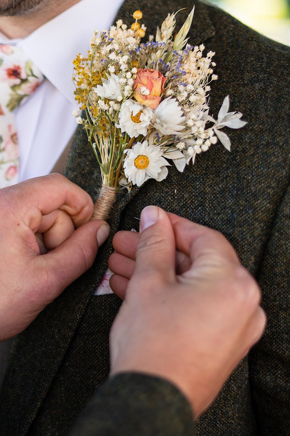 Hands pinning a rustic wildflower boutonniere with daisies onto a dark tweed suit jacket at a wedding.