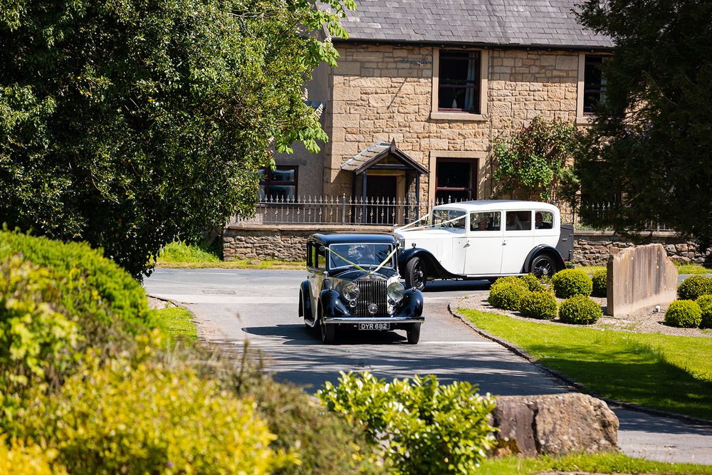 Two vintage wedding cars drive up a tree-lined driveway past a stone country house on a sunny day.