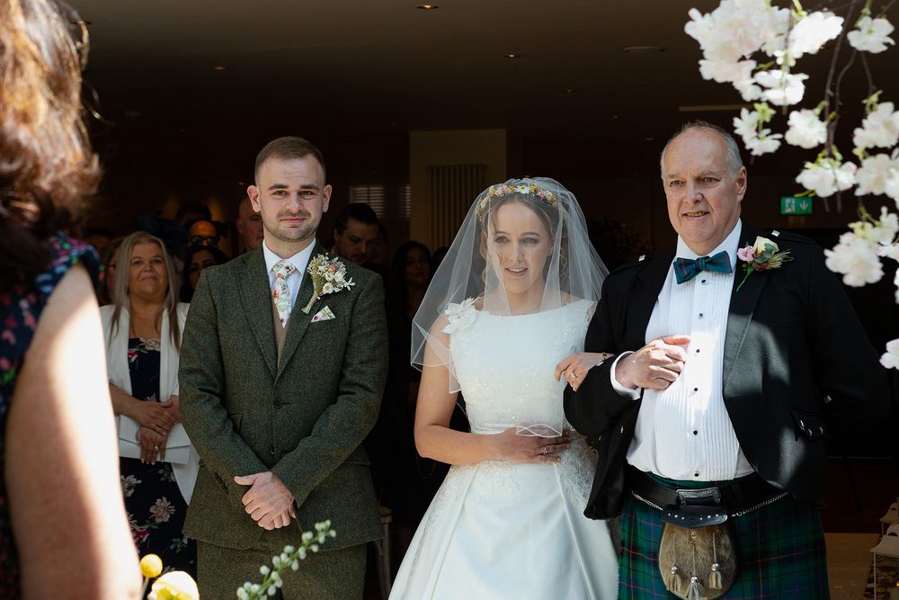 Bride in white gown walks down aisle with father in tartan kilt beside groom in green suit at wedding ceremony.