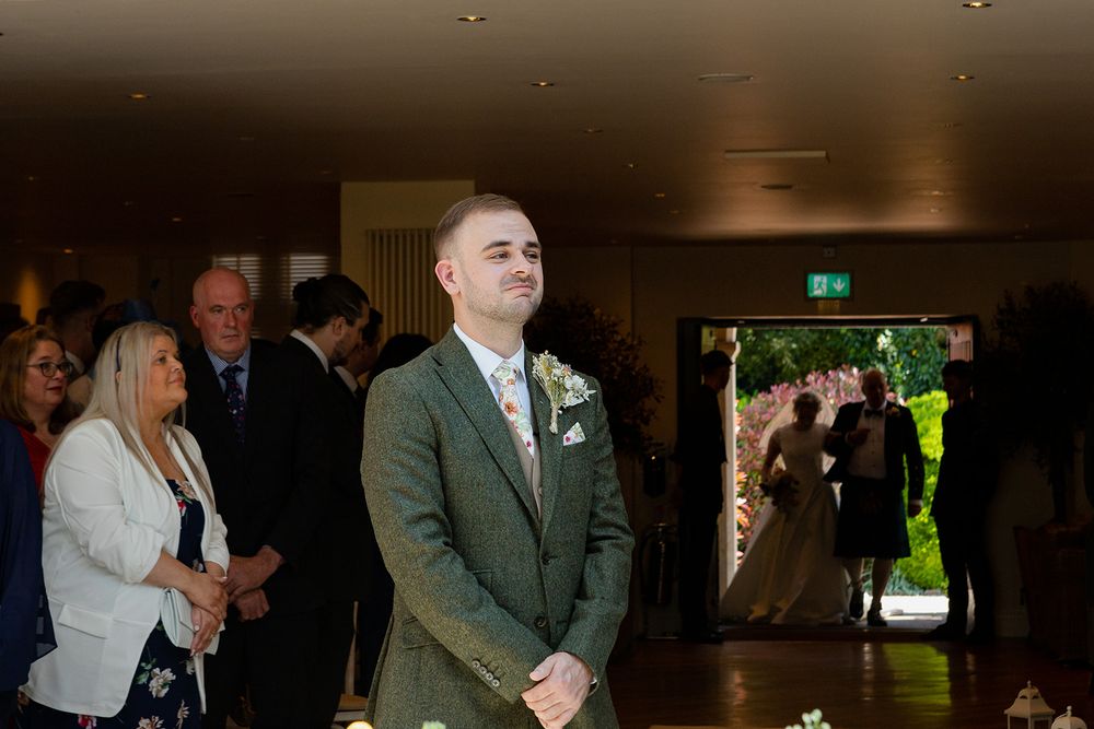 Groom in green tweed suit smiling at wedding ceremony altar as bride and wedding party stand in background.