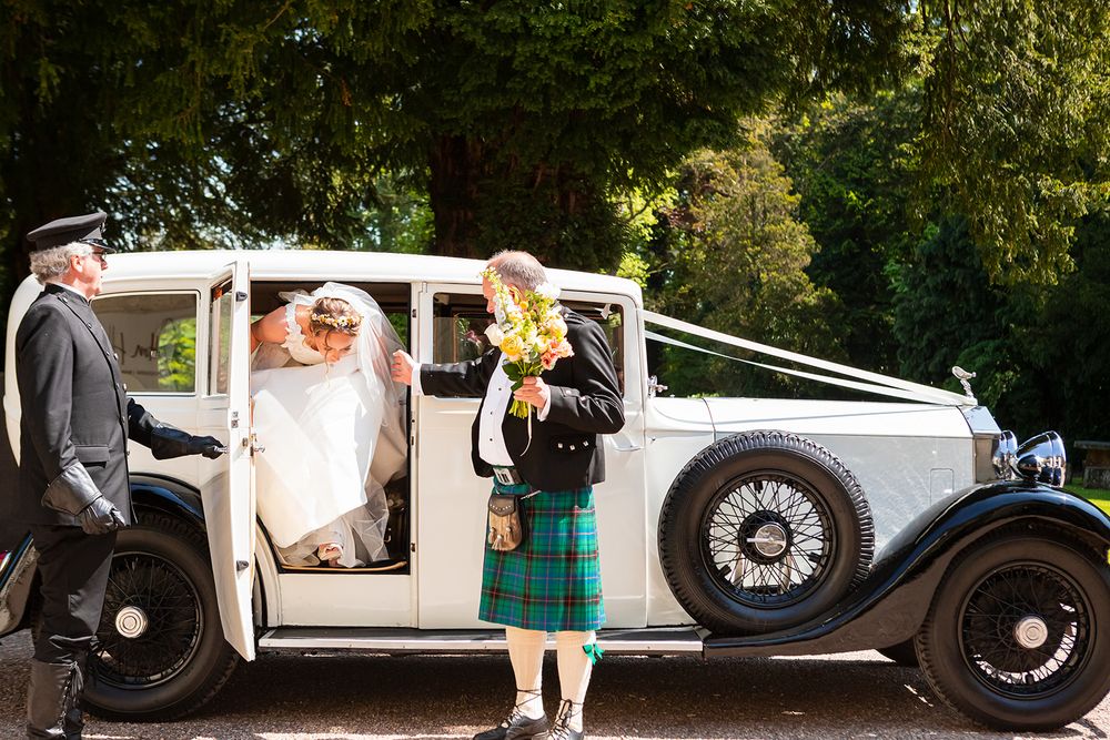 Bride in white gown exits vintage white wedding car as man in tartan kilt holds bouquet outside.