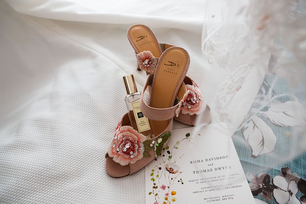 Blush pink floral embellished wedding heels with perfume bottle and invitation on white fabric.