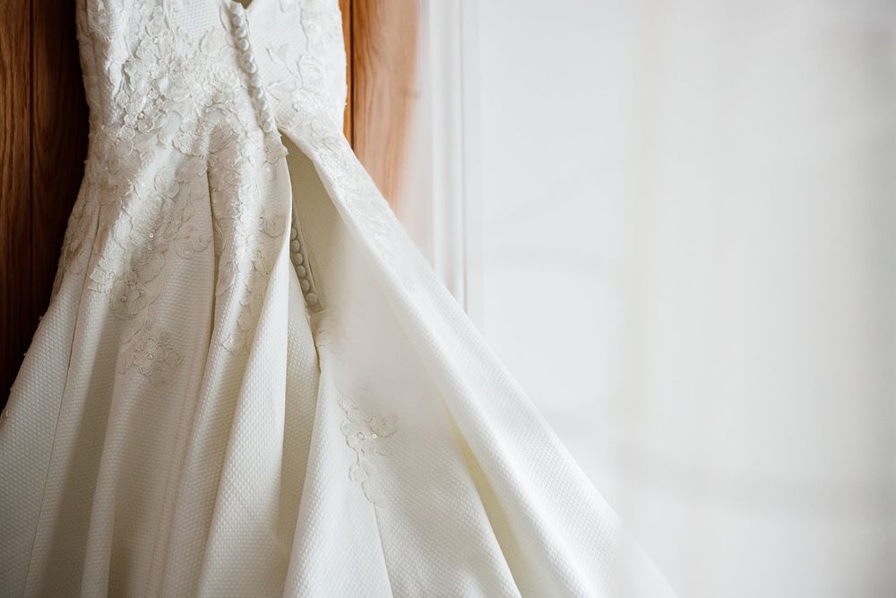 Elegant white lace wedding dress hanging by a window, showcasing intricate embroidery and a flowing satin skirt.