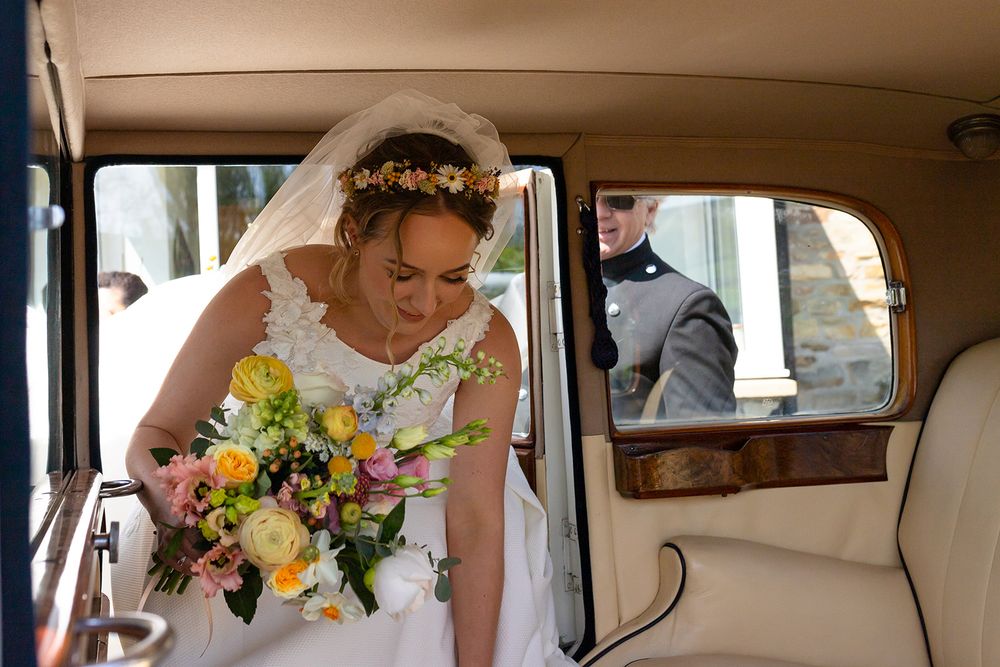 Bride in white gown holding colorful bouquet sits in vintage car, wearing floral crown and veil on wedding day.