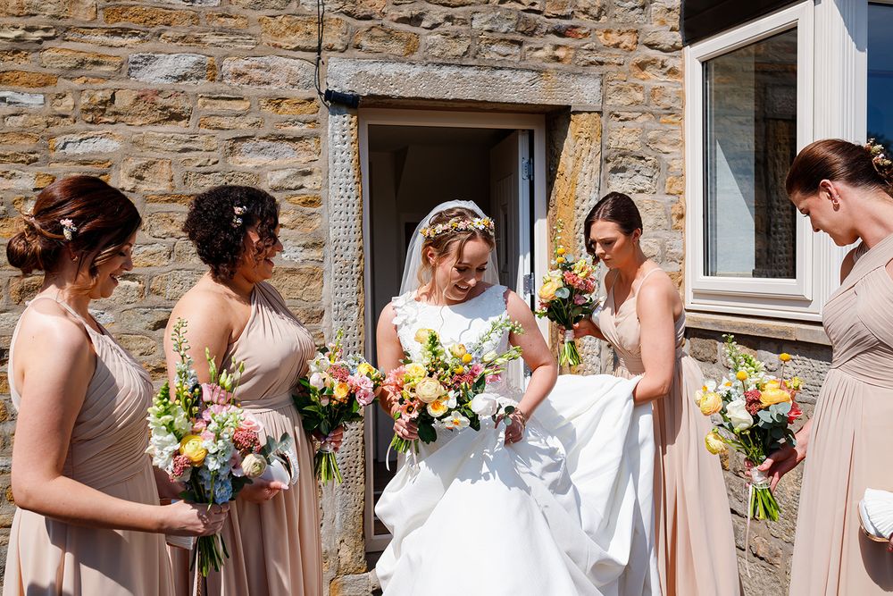 Bride in white gown with bridesmaids in champagne dresses holding colorful bouquets outside a stone building.
