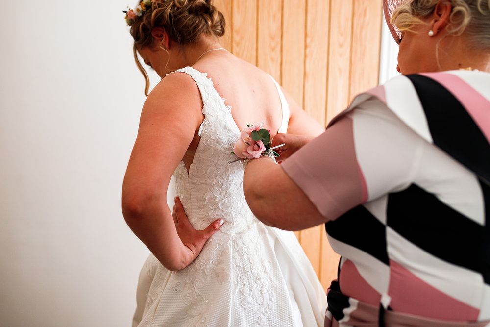 A woman helps a bride fasten the back of her white lace wedding dress before the ceremony.