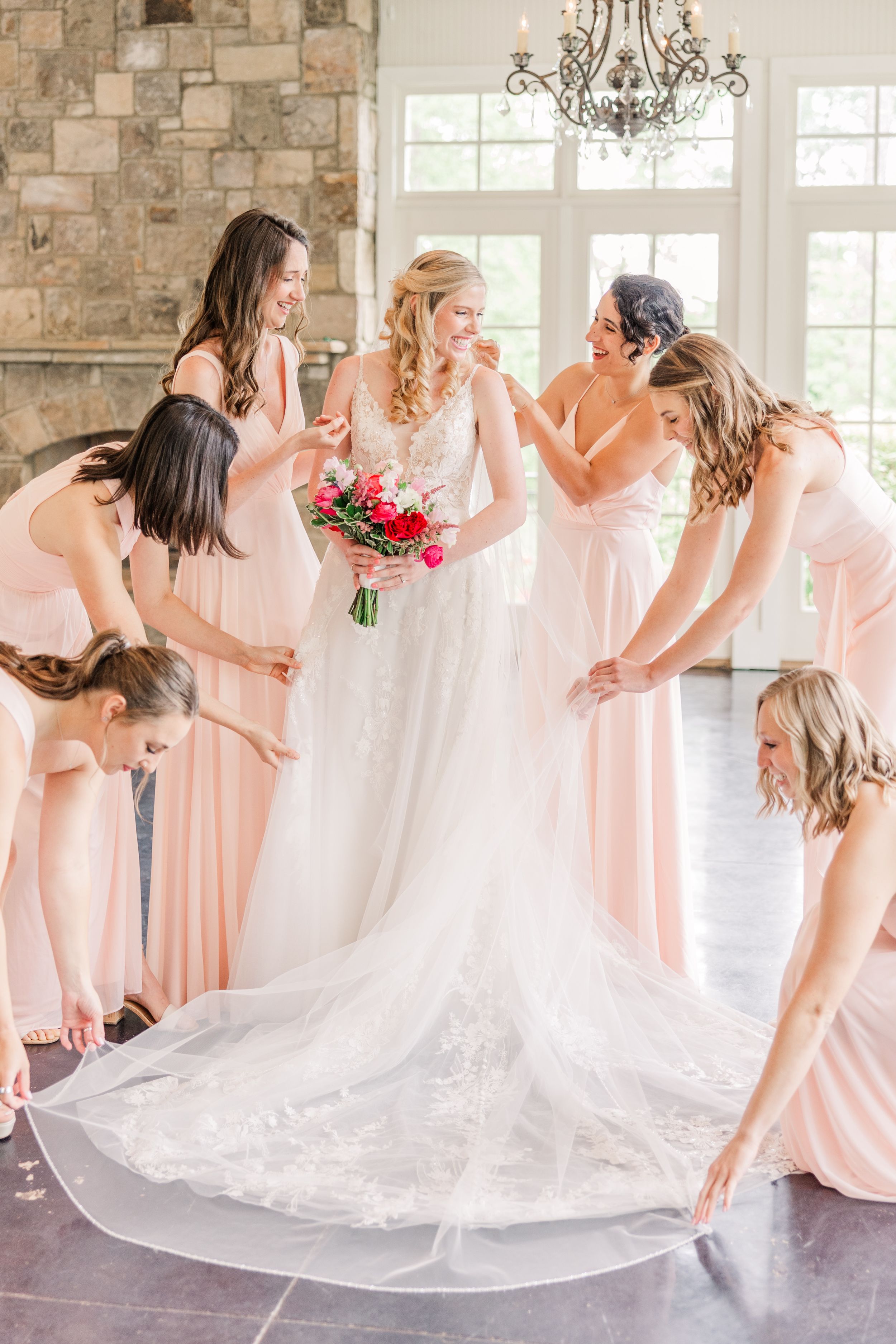 Bridesmaids in pink dresses gather around a bride, helping with her wedding gown.