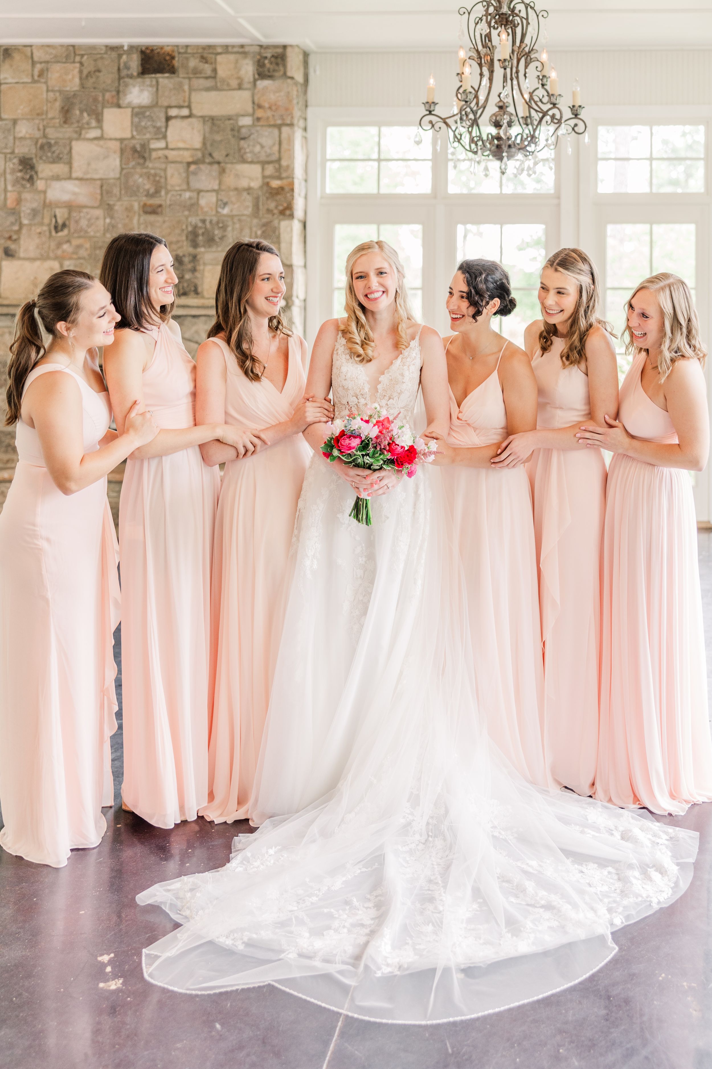 A bridal party in blush pink dresses poses together with the bride in her flowing white gown.