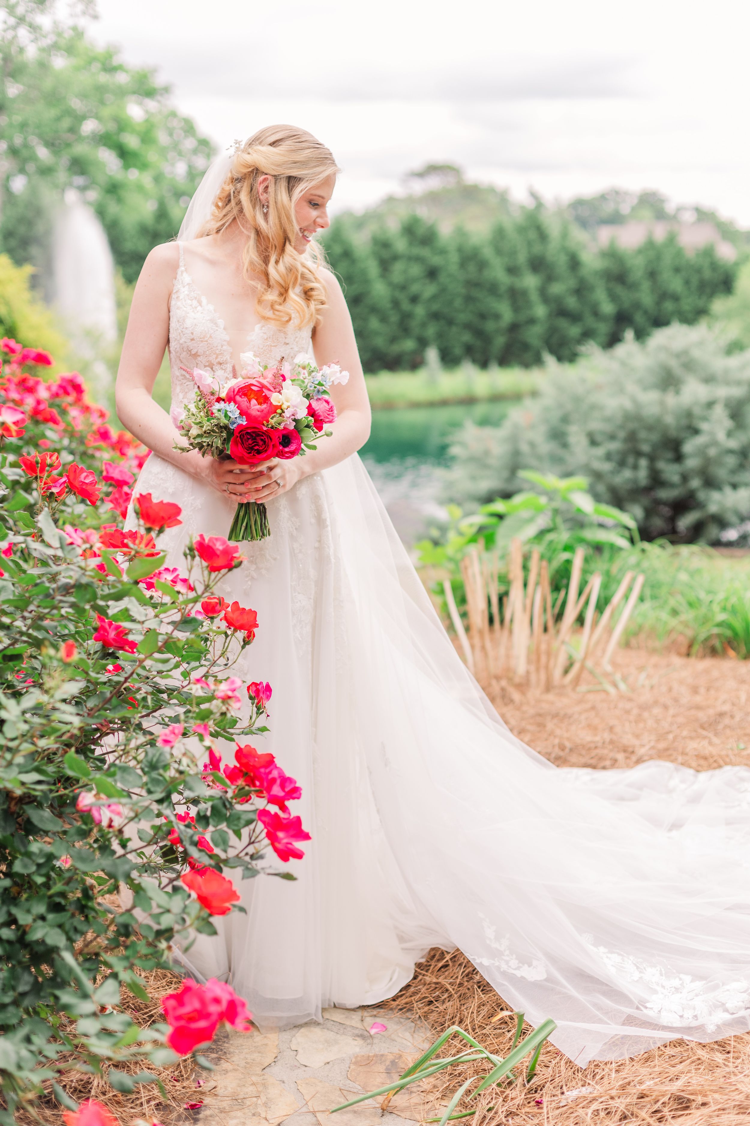 Bride in flowing white dress holds red bouquet next to rose bush in garden setting.