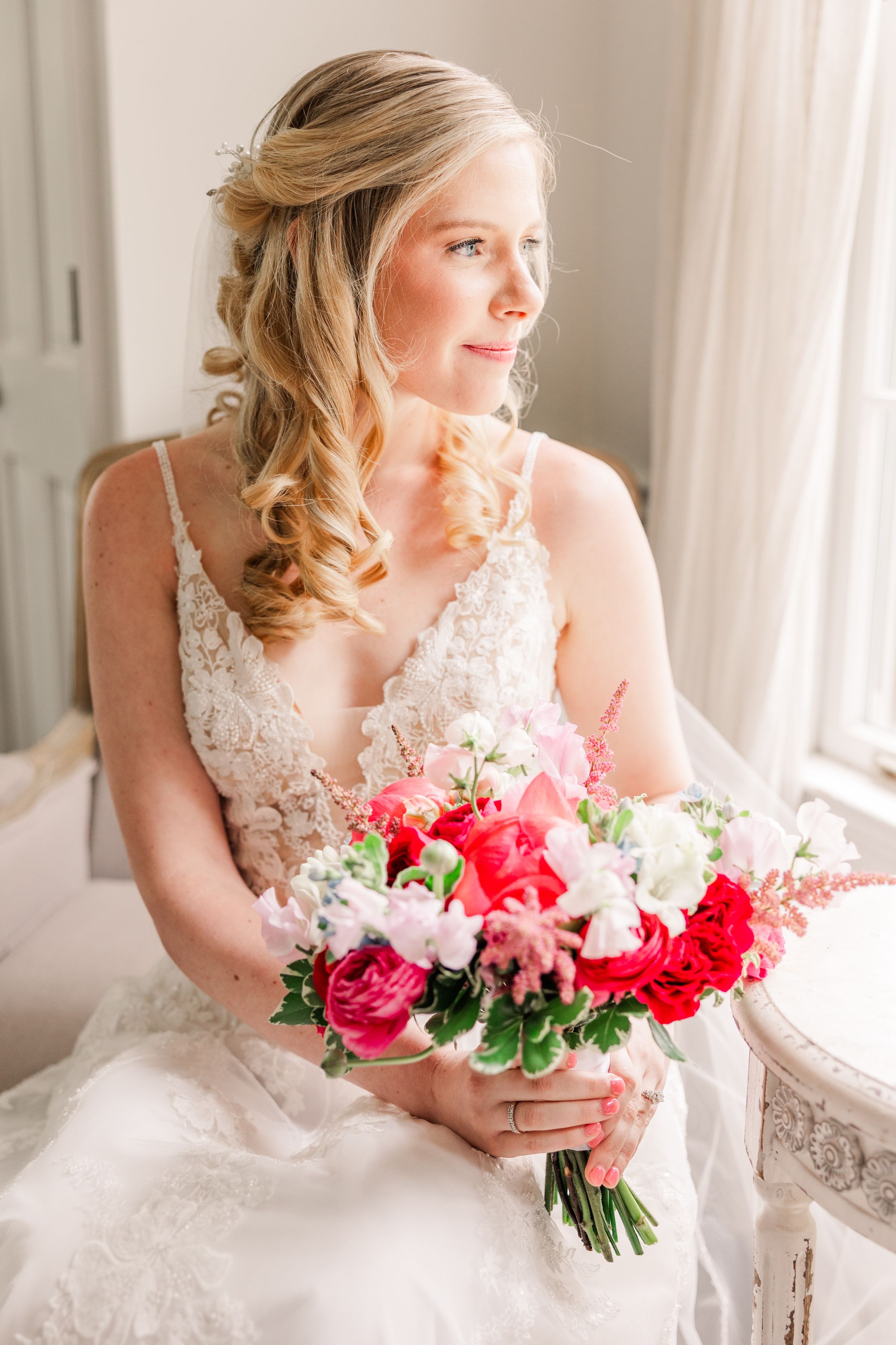 A bride in lace holds a vibrant bouquet of red and pink roses by a window.