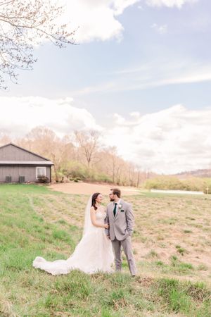Newlyweds pose together in an open field with rustic barn and spring trees in background.