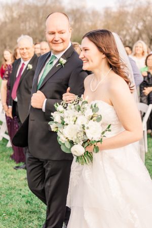 Wedding ceremony outdoors with guests seated on white chairs in a grassy field.